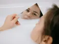 Kobieta w wannie z mlekiem ogląda w lustrze swoją twarz, zwracając uwagę na skórę pod oczami.