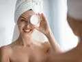 Uśmiechnięta kobieta z kremem na twarzy, ręcznikiem na głowie, pokazuje jak odbudować barierę hydrolipidową.