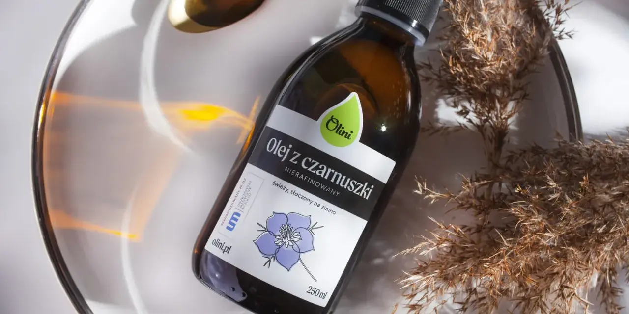 Olej z czarnuszki Olini, nierafinowany, tłoczony na zimno. Naturalny olejek na trądzik, zamknięty w szklanej butelce, otoczony suchymi trawami i złotą łyżeczką.