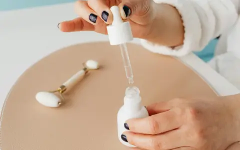 Dłoń z ciemnym lakierem do paznokci aplikuje serum z niacynamidem i kwasem salicylowym. Obok leży jadeitowy roller.