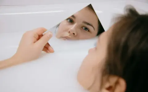 Kobieta w wannie z mlekiem ogląda w lustrze swoją twarz, zwracając uwagę na skórę pod oczami.