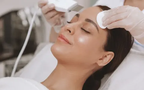 Kobieta przechodzi zabiegi nawilżające na twarz. Kosmetolog wykonuje ultradźwiękowy peeling, a druga dłoń delikatnie oczyszcza skórę.