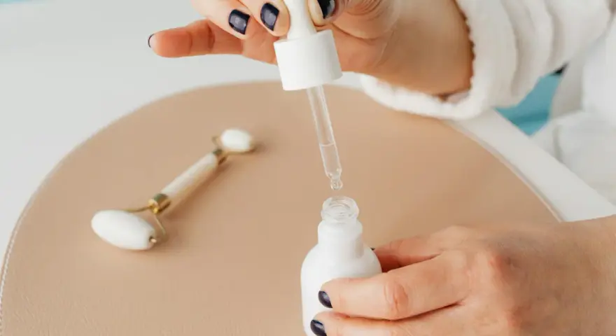 Dłoń z ciemnym lakierem do paznokci aplikuje serum z niacynamidem i kwasem salicylowym. Obok leży jadeitowy roller.