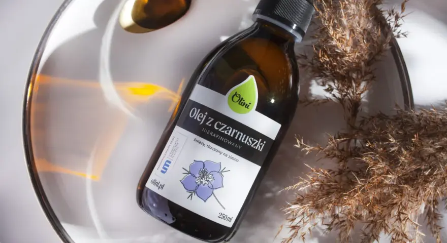 Olej z czarnuszki Olini, nierafinowany, tłoczony na zimno. Naturalny olejek na trądzik, zamknięty w szklanej butelce, otoczony suchymi trawami i złotą łyżeczką.